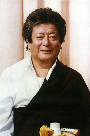 Thinley Norbu Rinpoche wearing a white shirt, red zen, pink curtain in background.jpg